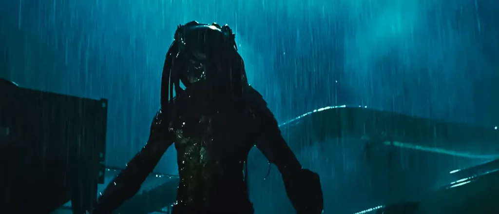 Ein Predator aus "Alien vs. Predator: Requiem" steht kampfbereit im Regen bei Nacht, beleuchtet von schwachem Licht, mit glühenden Augen und gezückten Waffen.